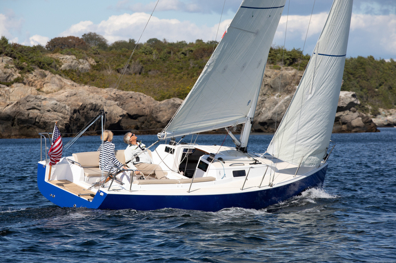 Sailboat with two people near rocky shore.