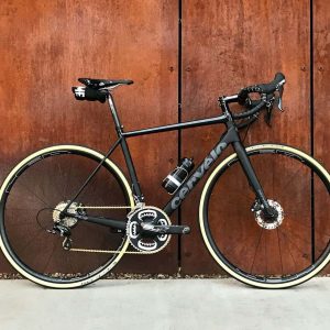 A sleek black road bike with tan tires leaning against a wooden wall.