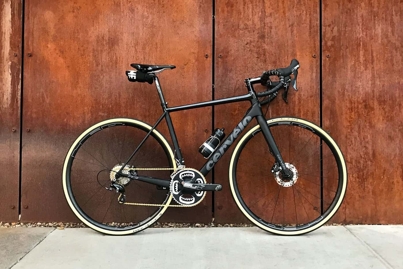 A sleek black road bike with tan tires leaning against a wooden wall.