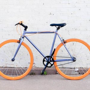 A stylish fixed-gear bicycle with bright orange wheels against a white brick wall.