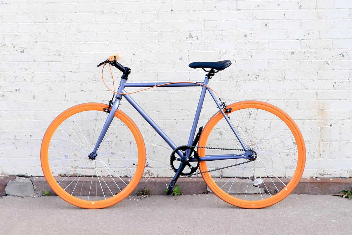 A stylish fixed-gear bicycle with bright orange wheels against a white brick wall.