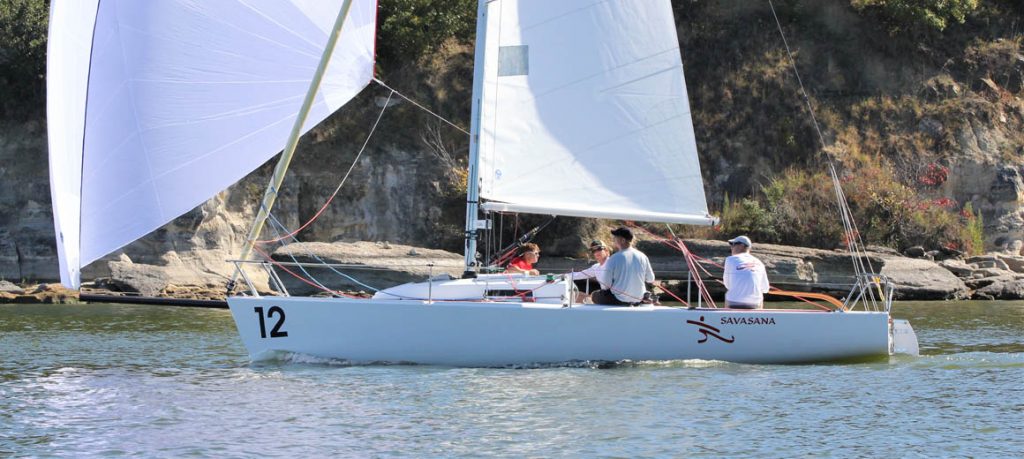 Person sailing a small white sailboat on calm water near a forested shore.