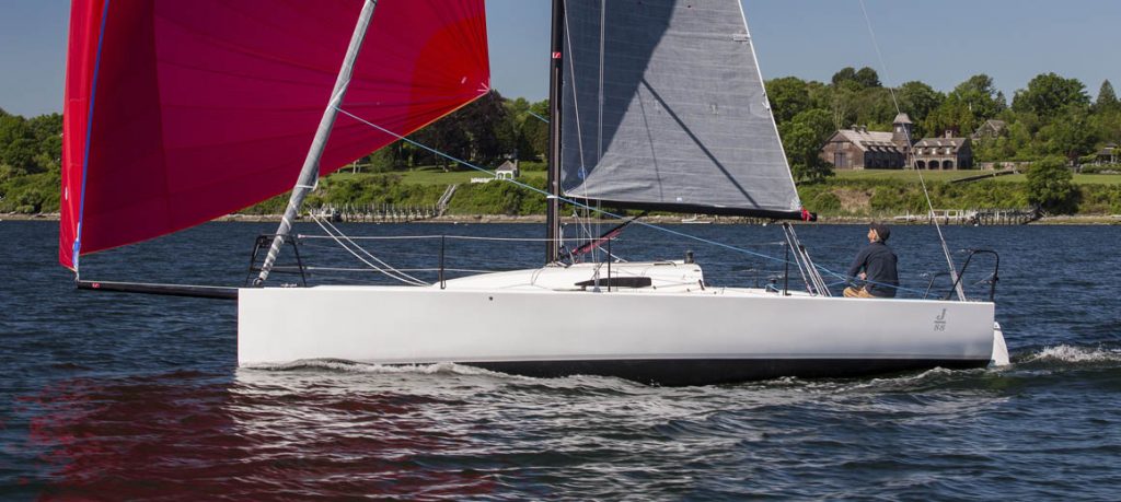 Sleek white sailboat with gray and red sails on calm water near green shore.