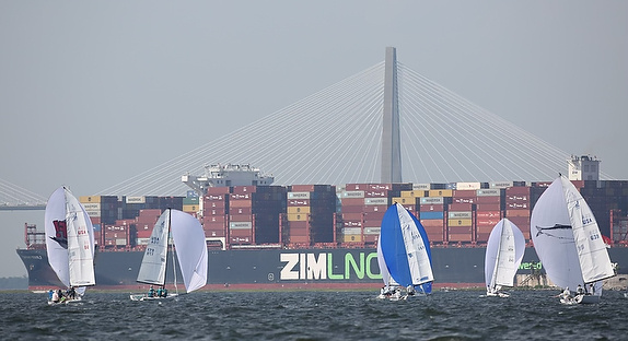 Sailboats race near a large cargo ship with containers and a bridge in the background.