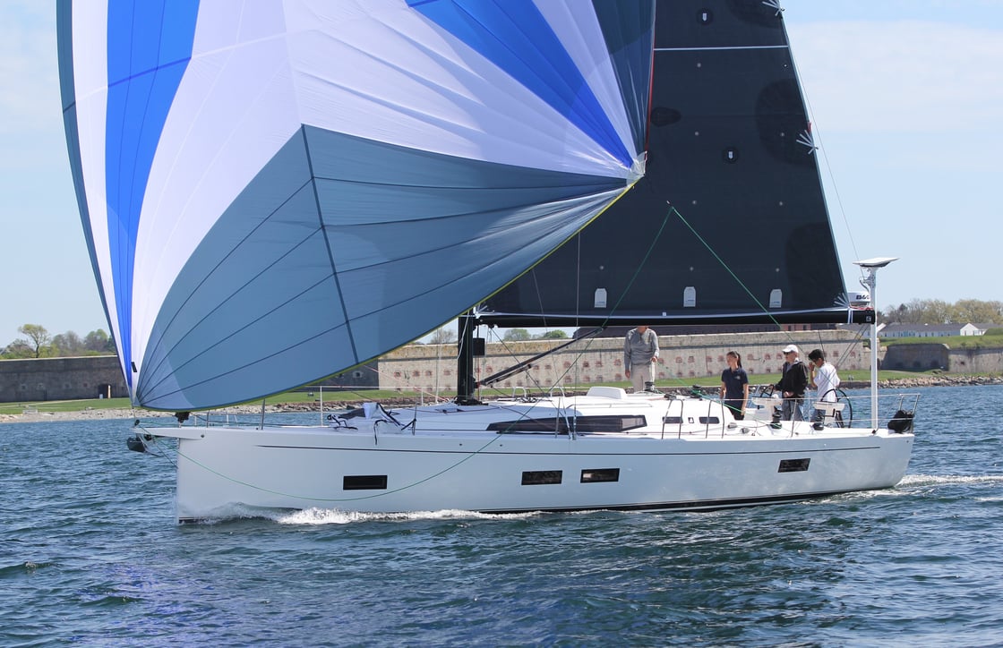Sailboat with blue and white spinnaker sails glides on calm water near a dock.