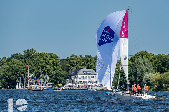 Sailboat with colorful spinnakers glides on a lake near a waterfront house.