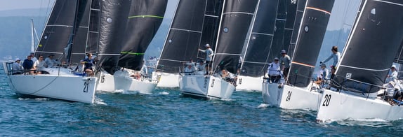 Three sailboats racing on clear blue water with sailors maneuvering sails.